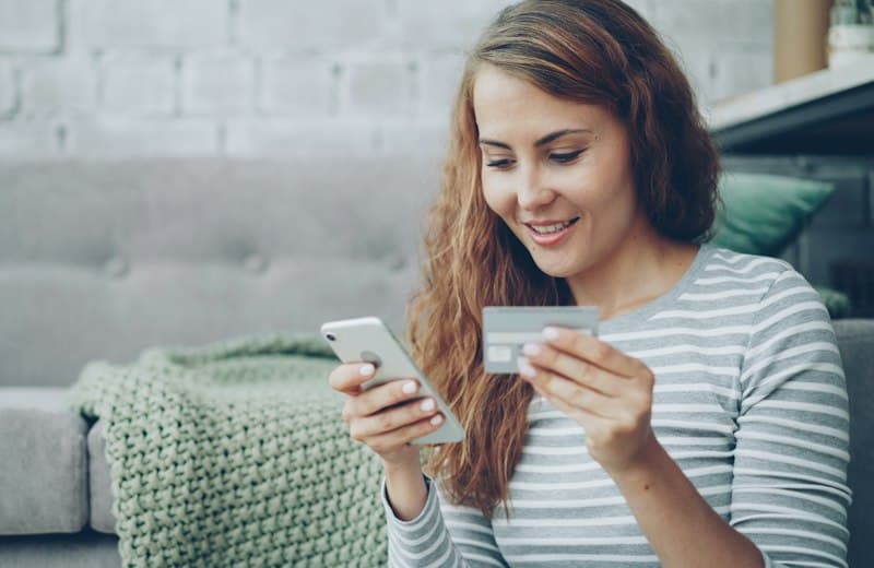 Mujer sonriendo mientras paga en línea con su celular y una tarjeta